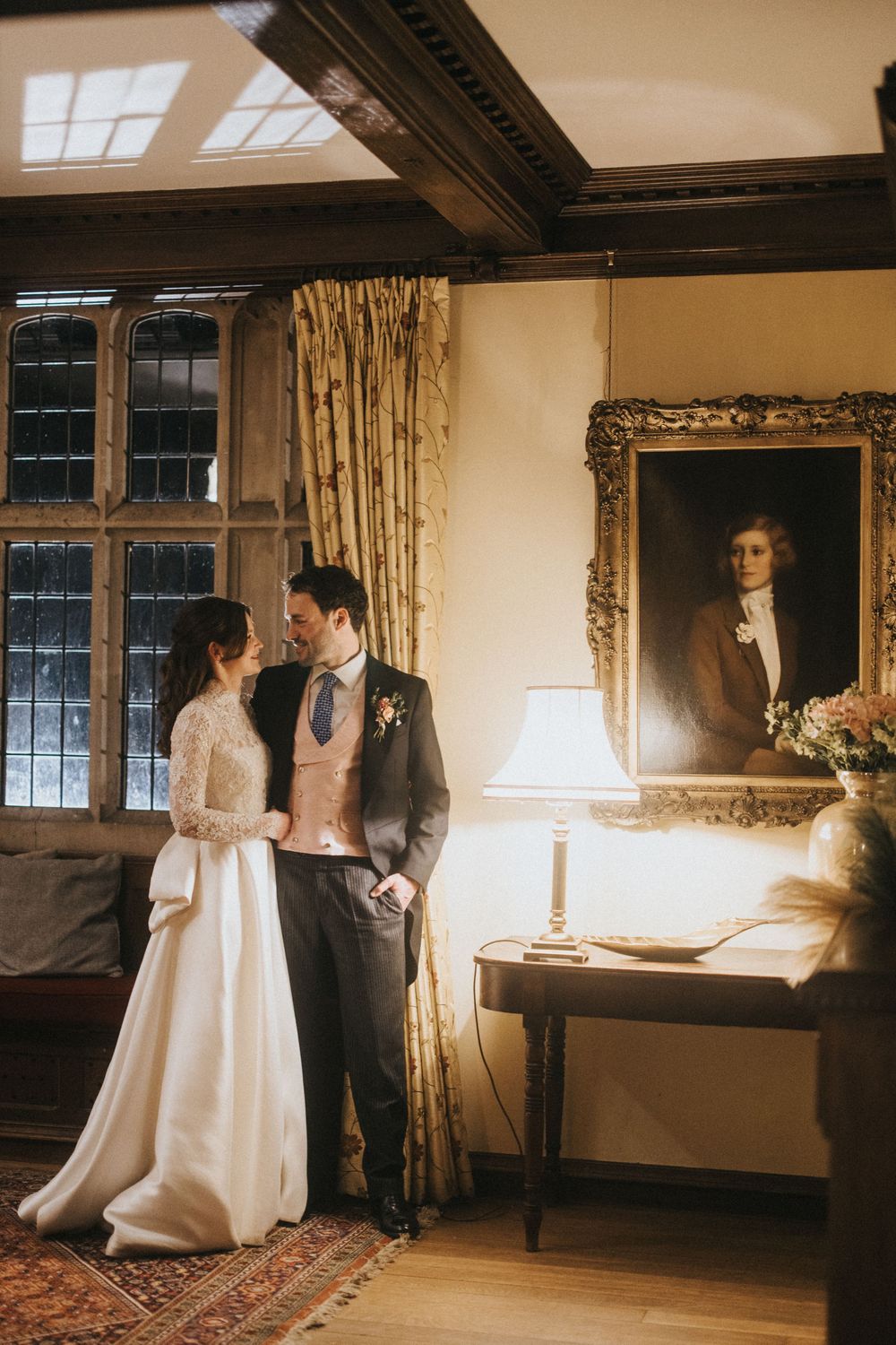 A romantic wedding couple poses in an elegant vintage room with ornate decor and gilt-framed portrait.