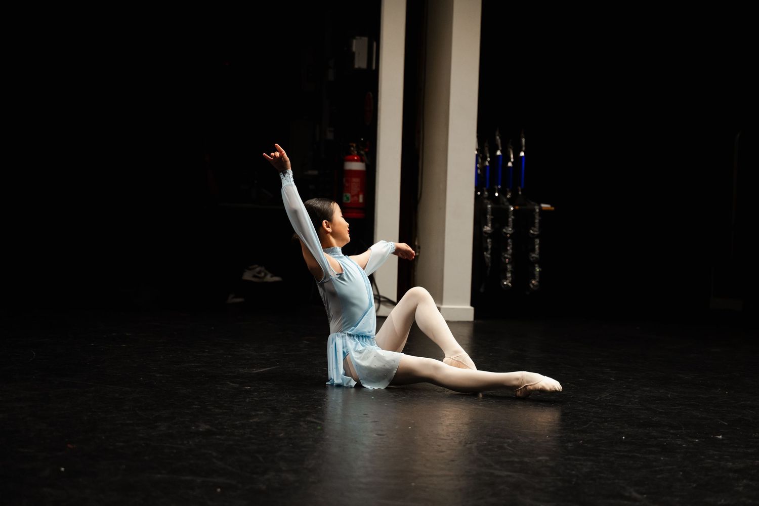 Ballet dancer in light blue leotard performs graceful floor pose on dark stage with elegant arm extension upward.