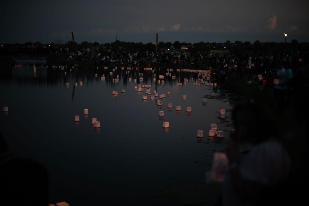 Glowing lanterns float across a dark lake at night as crowds gather along the shore during a festival celebration.