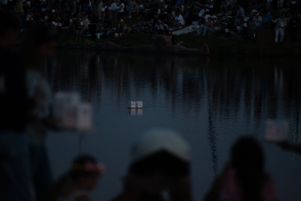 Silhouetted figures watch illuminated windows reflecting on dark water at dusk.