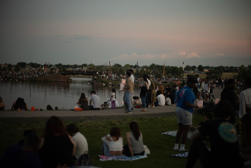 People gather on grassy hill at sunset to watch the evening sky over a city skyline in the distance.