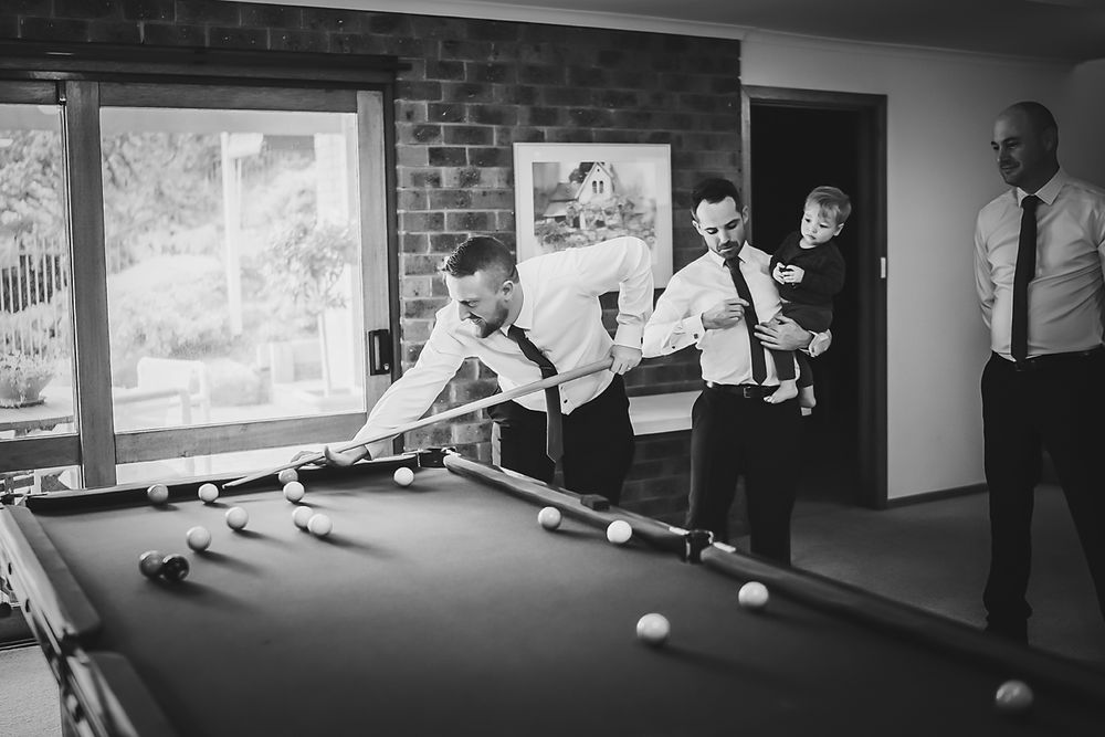 Black and white photo of people playing pool in a game room with brick walls and framed artwork.