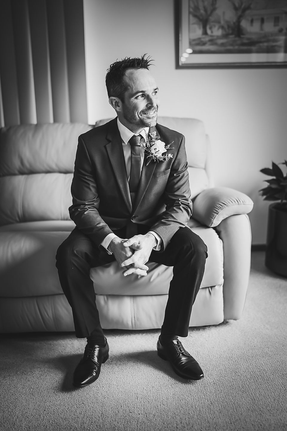 Black and white portrait of groom in formal suit sitting on light colored couch with boutonniere and bright smile.