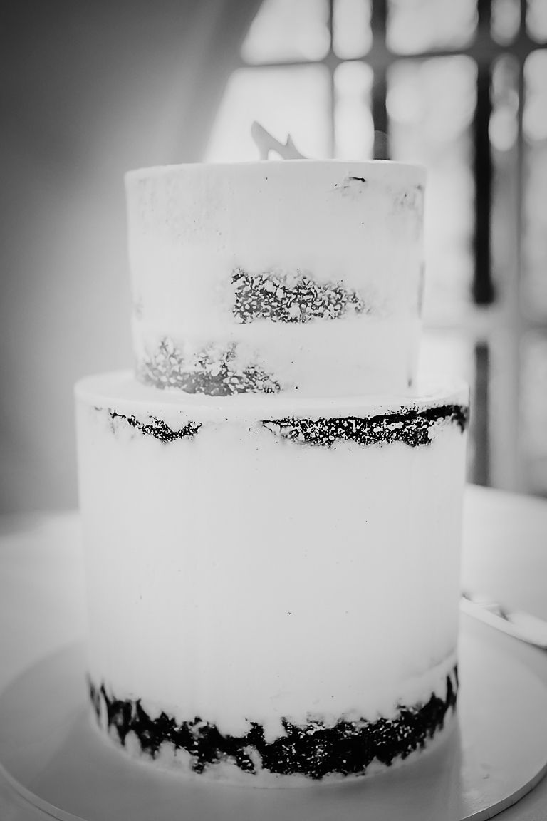 A black and white closeup photo of a frosted naked wedding cake with dark layers visible through semi-transparent icing.