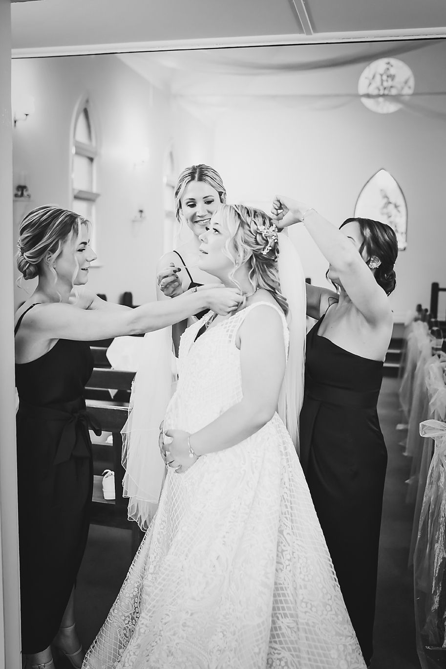 A candid black and white photo of bridal preparations showing a bride in a white gown with attendants in dark dresses.