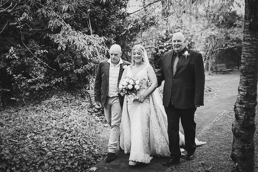 A black and white wedding party walks along a tree-lined path during an outdoor celebration ceremony.