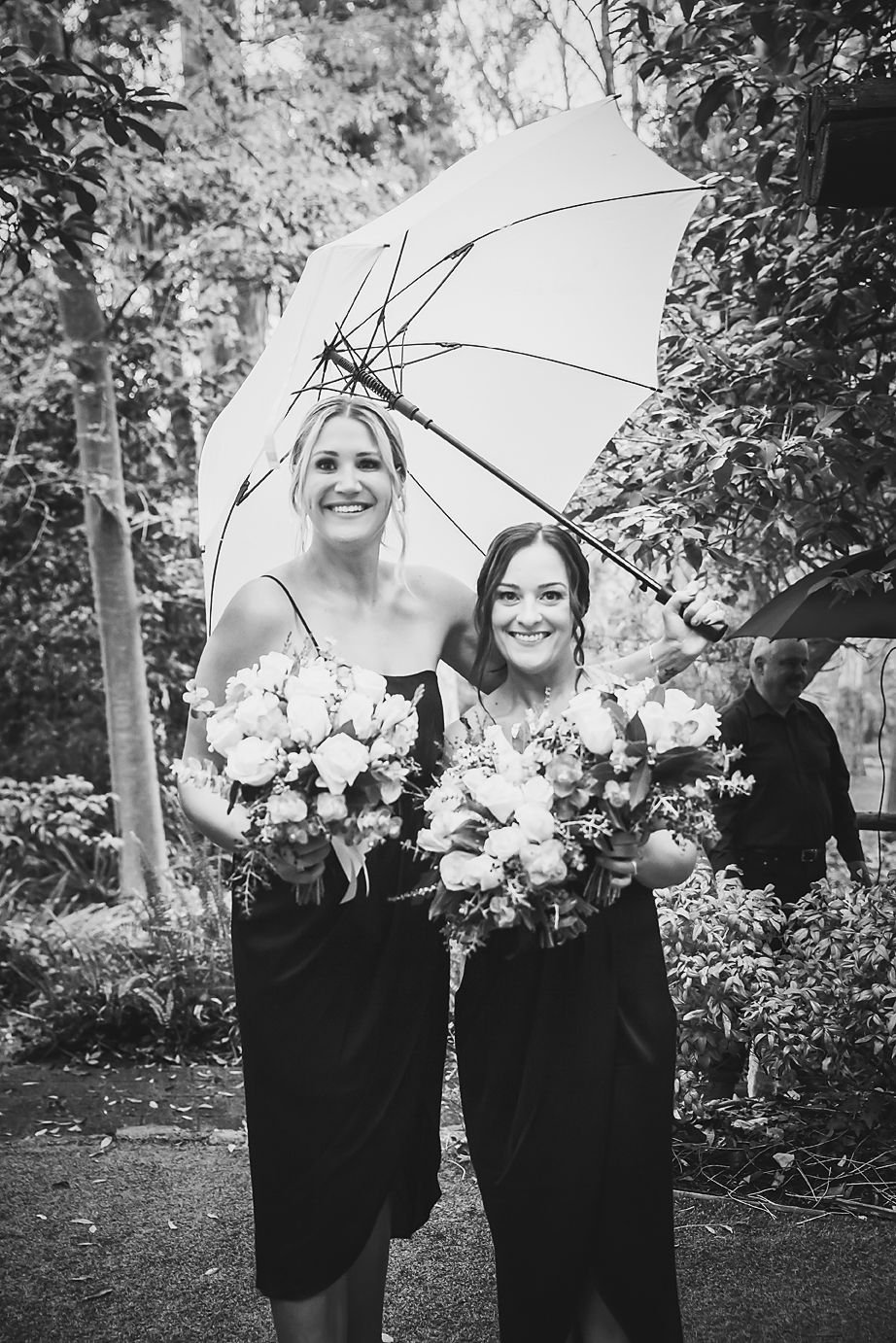 Black and white photo of bridesmaids in dark dresses holding floral bouquets under a white umbrella in a garden setting.