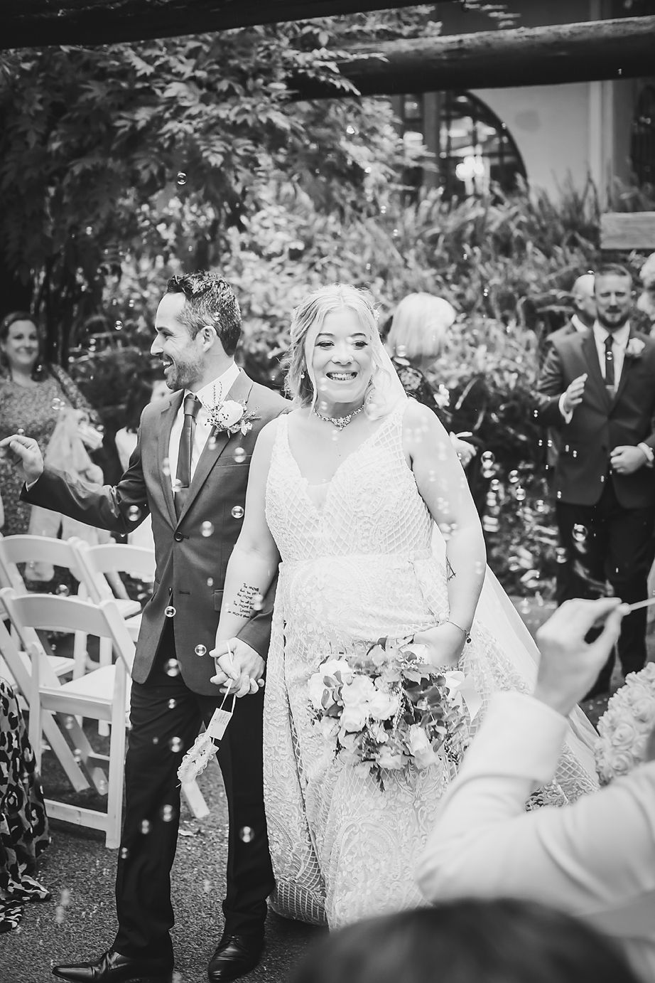 Black and white photo of a joyful couple celebrating at an outdoor garden wedding ceremony with guests tossing petals.