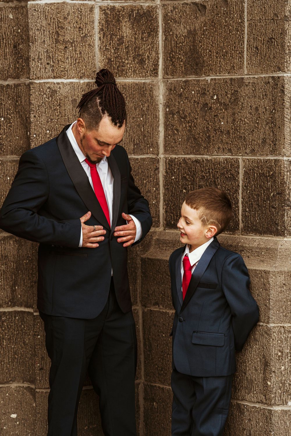Adult and child in matching black suits with red ties stand against stone wall while adjusting their outfits.