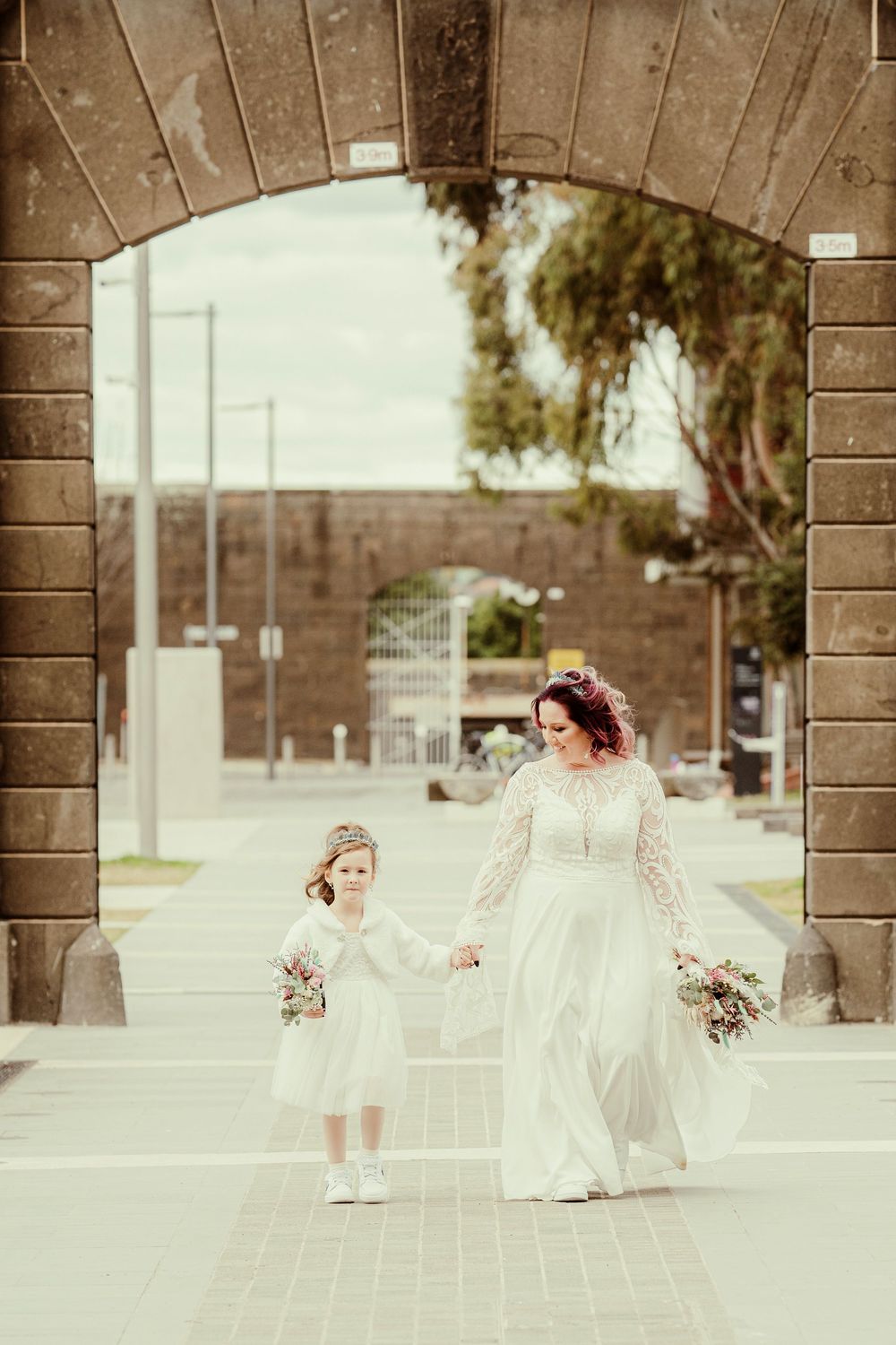 Two people in white dresses and flower bouquets stand under a stone archway in a vintage-style photograph.