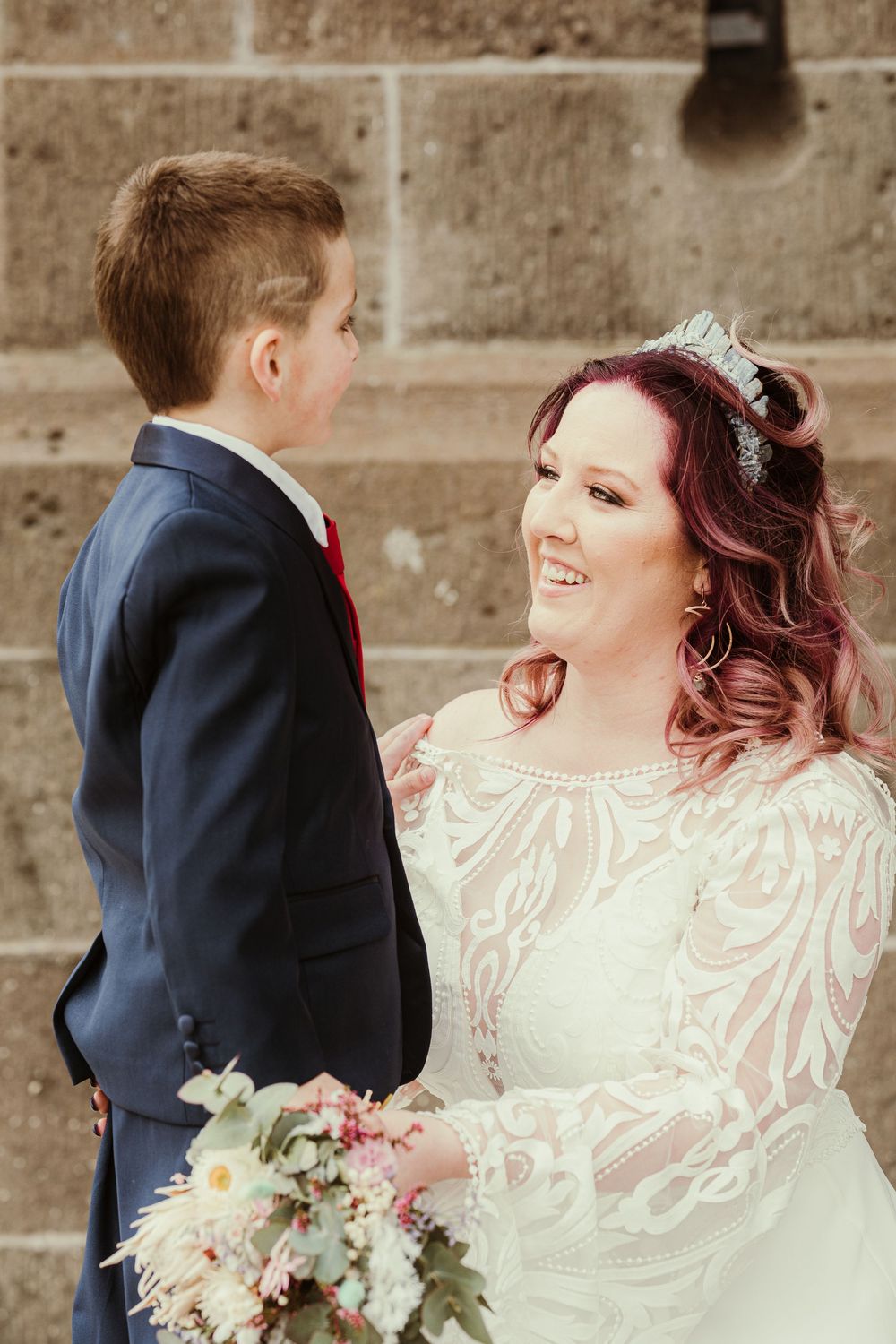 Bridal couple in navy suit and lace dress share a romantic moment against a stone wall with pink and white bouquet.