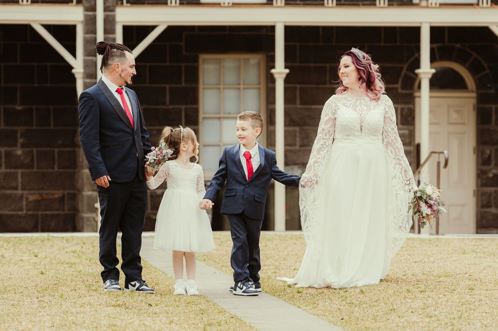 A sunny outdoor family wedding photo with loved ones standing together on a grassy lawn in front of a rustic brick building.