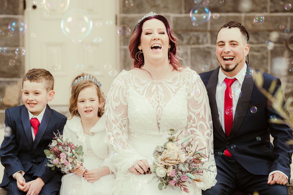 Wedding party laughs together in formal attire against a stone wall backdrop with vibrant red accents.