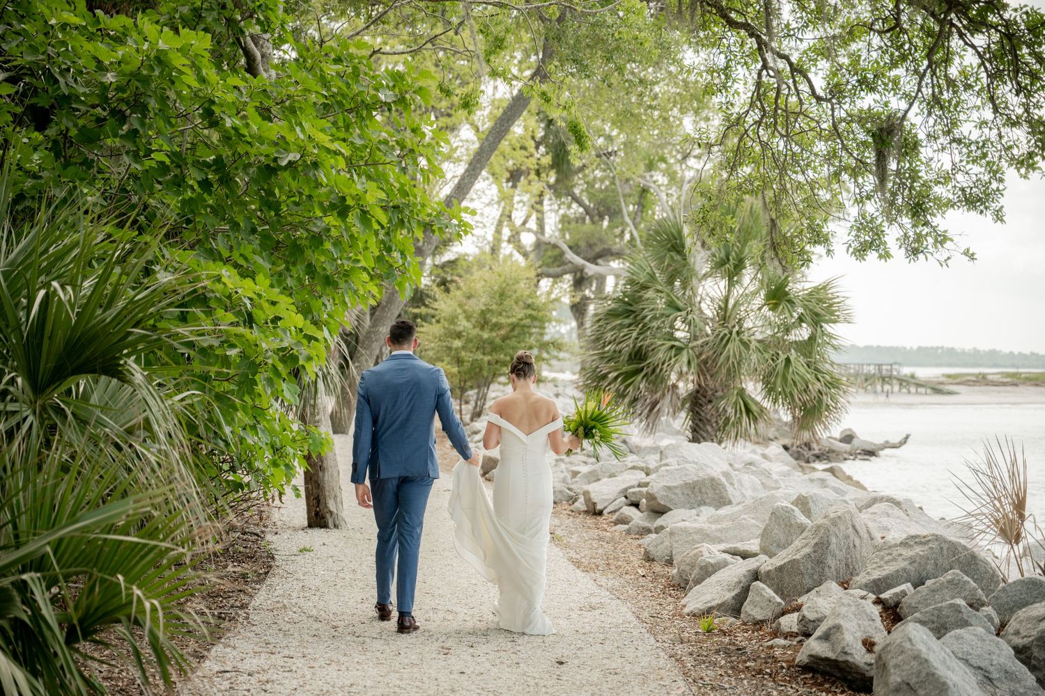 A couple in wedding attire walks down a palm-lined path along a rocky shoreline on a sunny day.