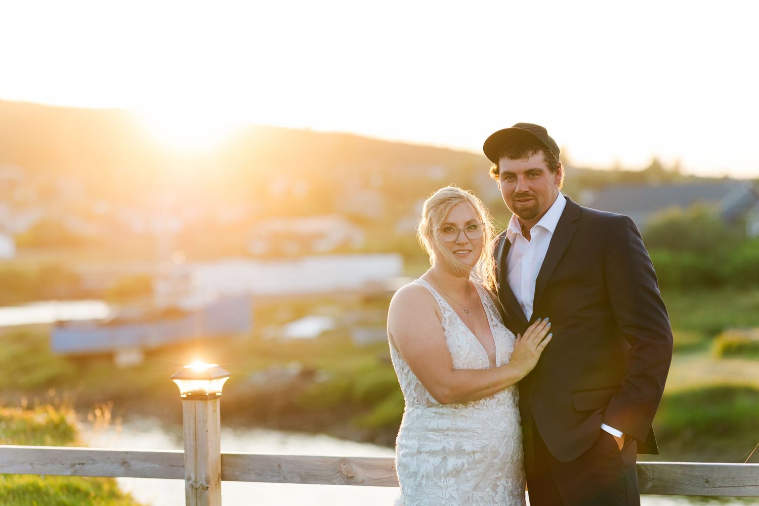 A bride and groom pose together on a deck during sunset with golden light illuminating the background.