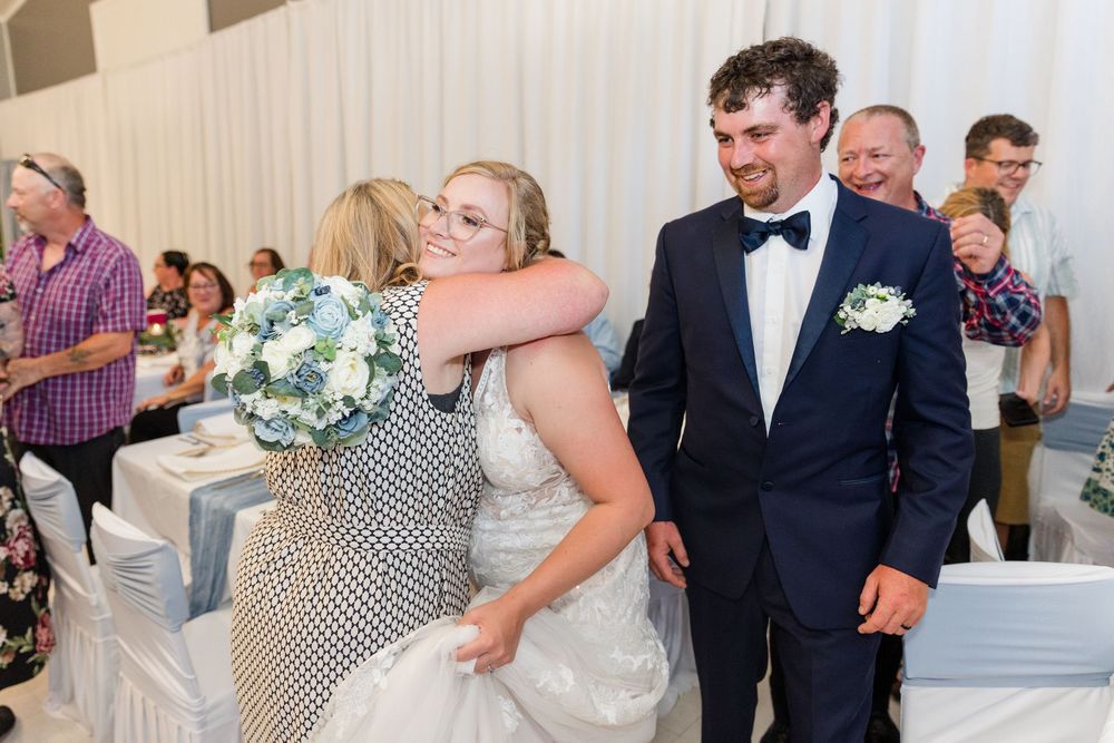 A wedding party embraces while celebrating during a reception in a bright white-decorated event space.
