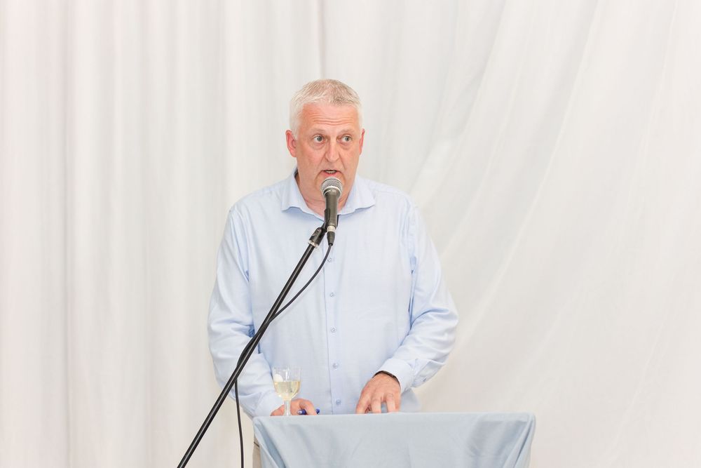 Speaker standing at podium wearing light blue dress shirt against white backdrop during presentation.