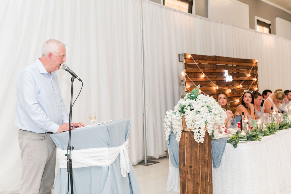 A speaker addresses wedding guests at a rustic reception with wooden decor and white floral arrangements.
