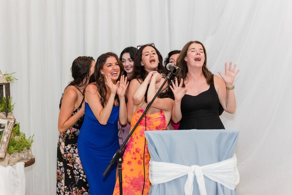 A group of friends in formal dresses laugh and react joyfully at a wedding reception with a white draped backdrop.