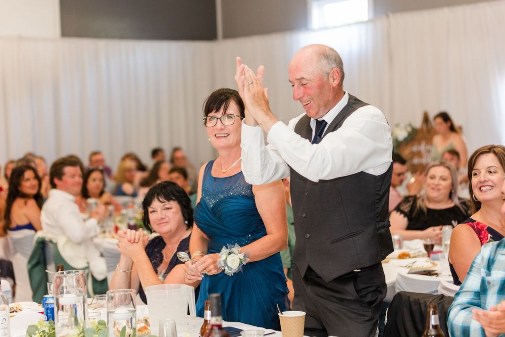 Reception guests applauding during wedding celebration in bright indoor venue with decorative greenery.