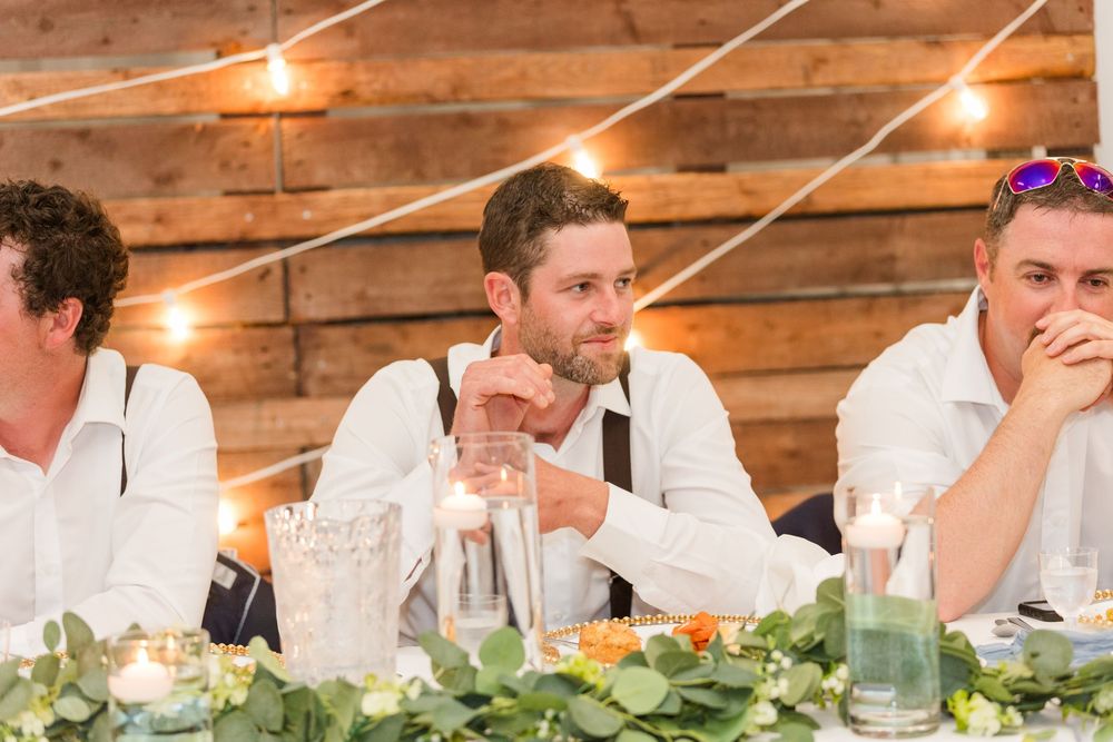 Groomsmen in white shirts and suspenders enjoying drinks at rustic wooden reception venue with string lights.
