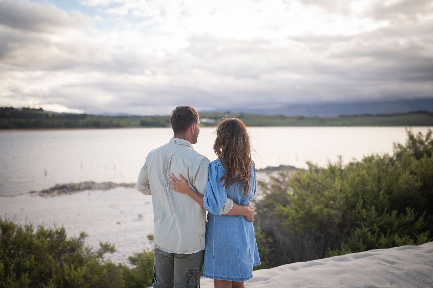 Two people stand at a scenic overlook taking in views of a tranquil lake surrounded by green hills under cloudy skies.