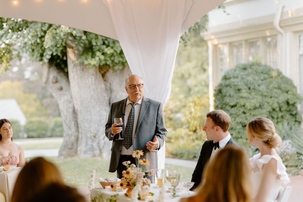 A person gives a speech during an elegant outdoor wedding reception under a white tent with string lights.