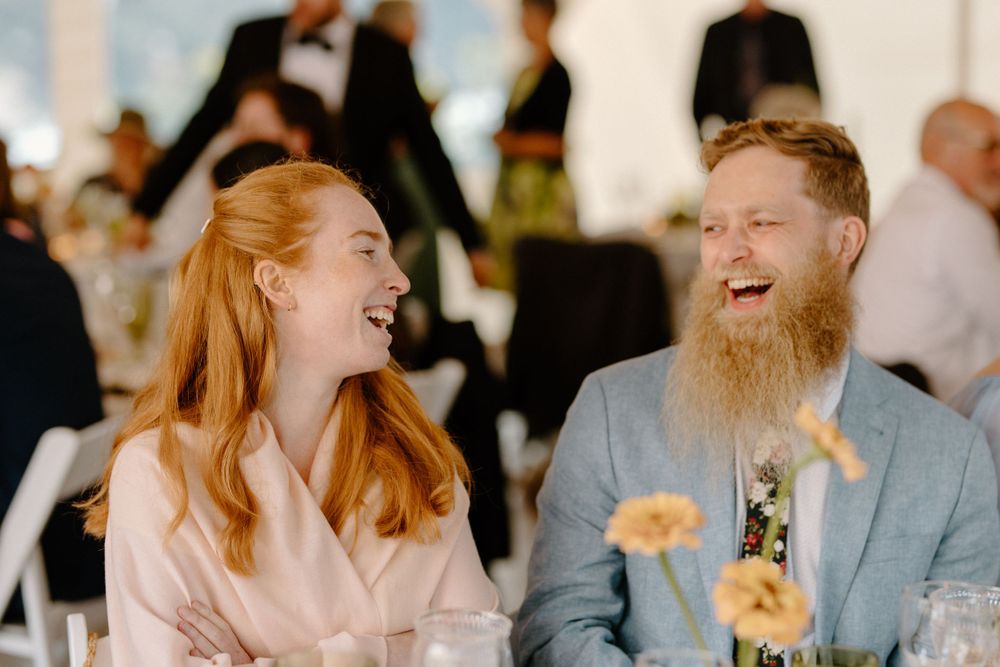 A couple shares a genuine laugh together at a wedding reception, with peach flowers decorating their table.