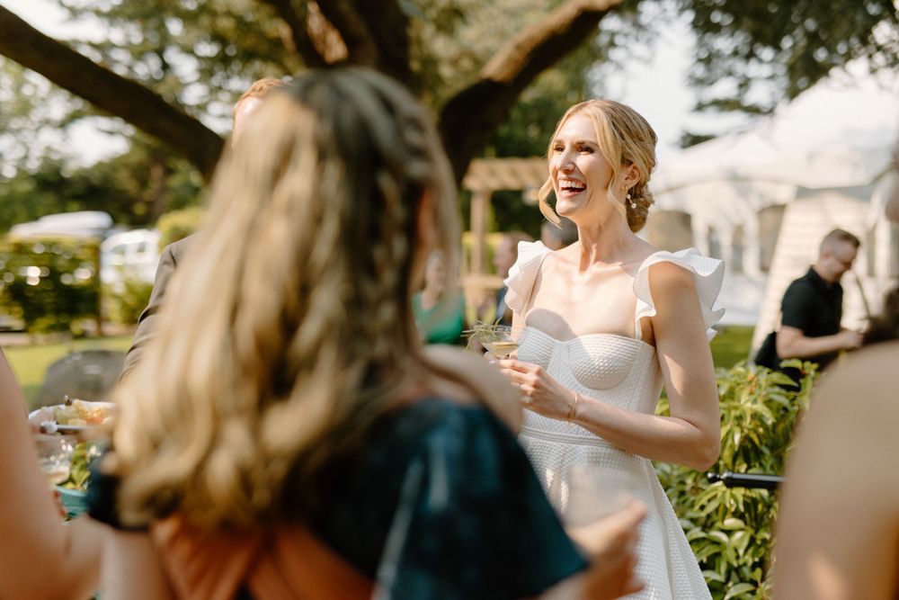 A bride in a white dress laughs during an outdoor garden reception while talking with guests.