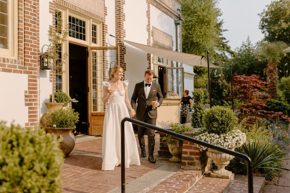 A couple stands together on an elegant brick porch with lush greenery and landscaping on a sunny autumn day.