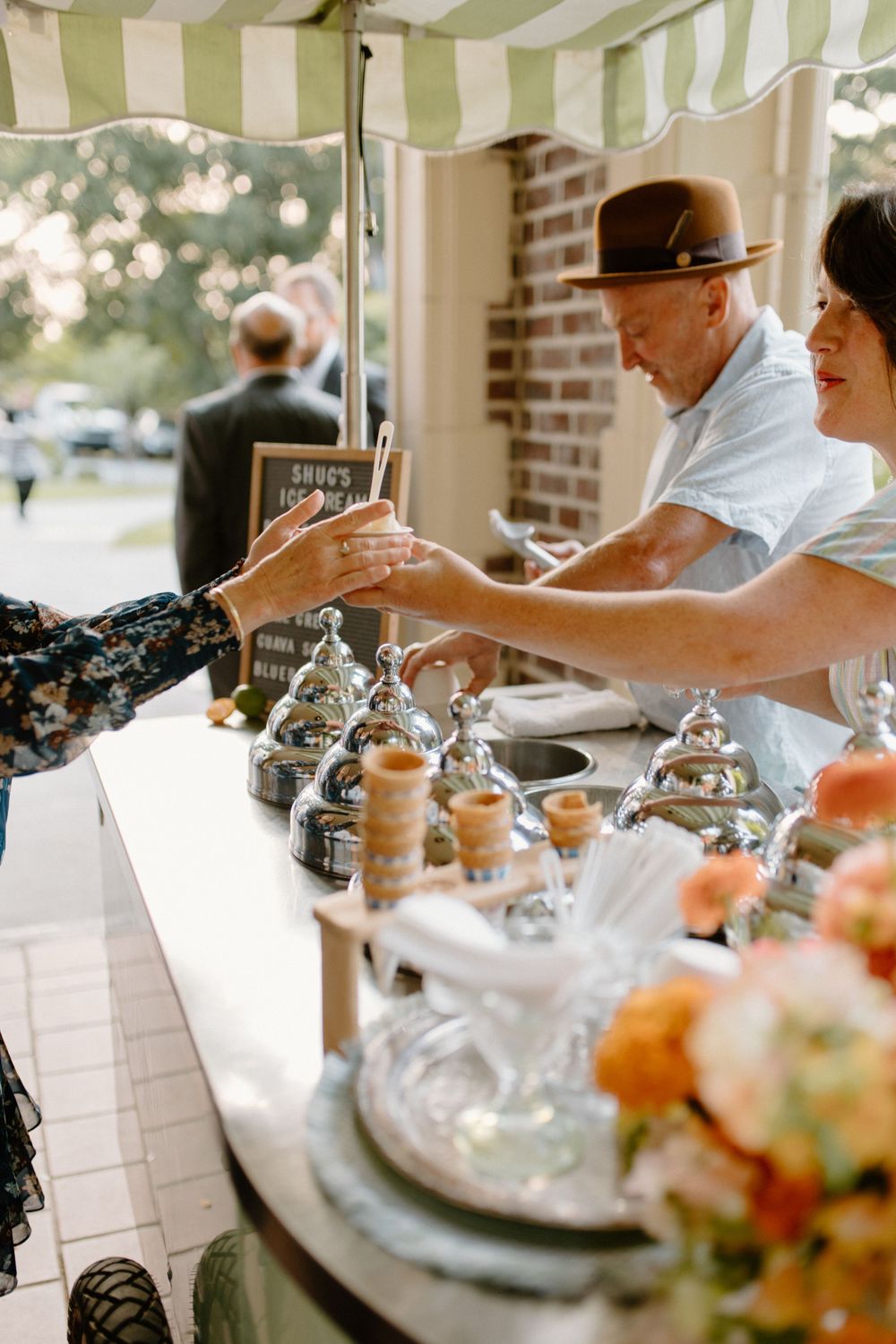 Guests and staff interact at an outdoor catering station with desserts and drinks under a striped awning.