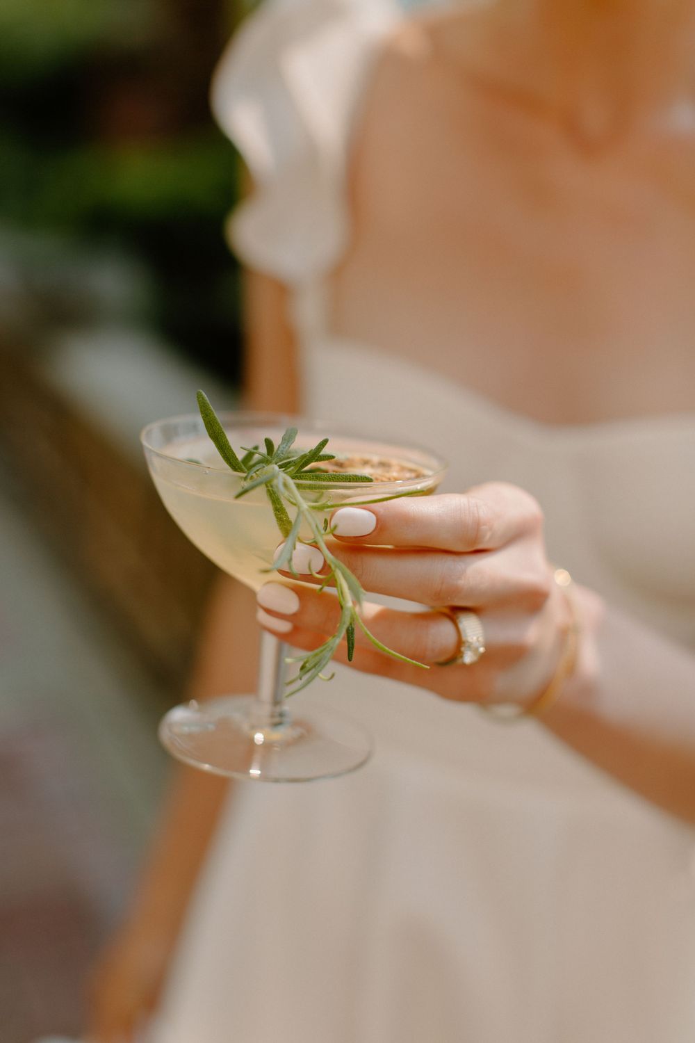 Elegant cocktail garnished with fresh rosemary held by manicured hands in white against a blurred background.