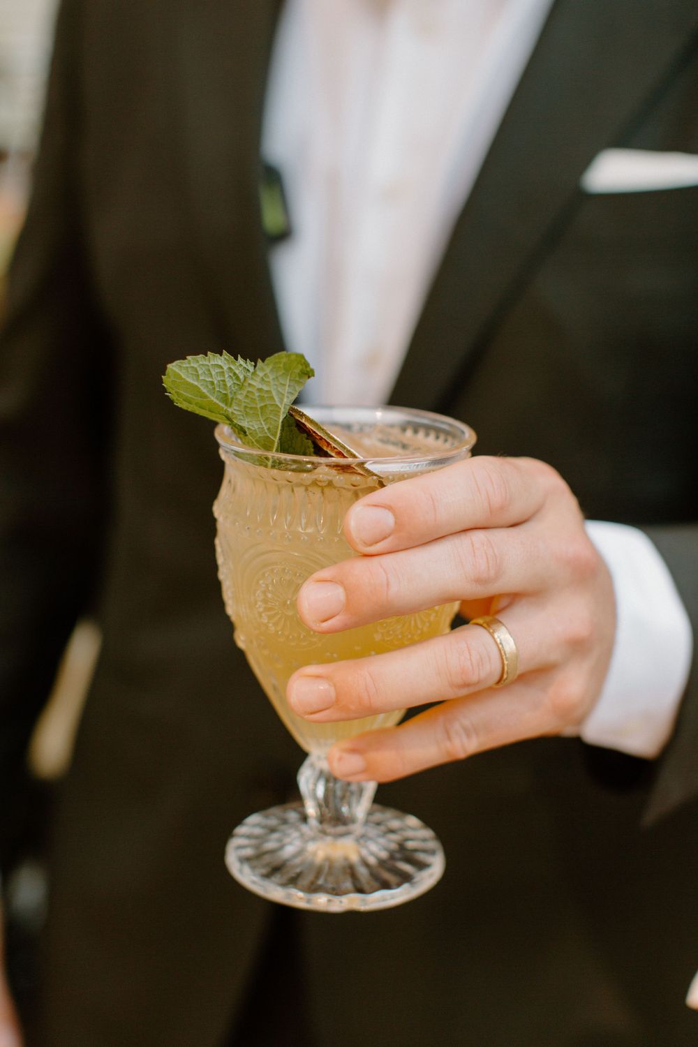 A hand wearing a suit sleeve and wedding ring holds a garnished cocktail in a crystal glass with a mint leaf.