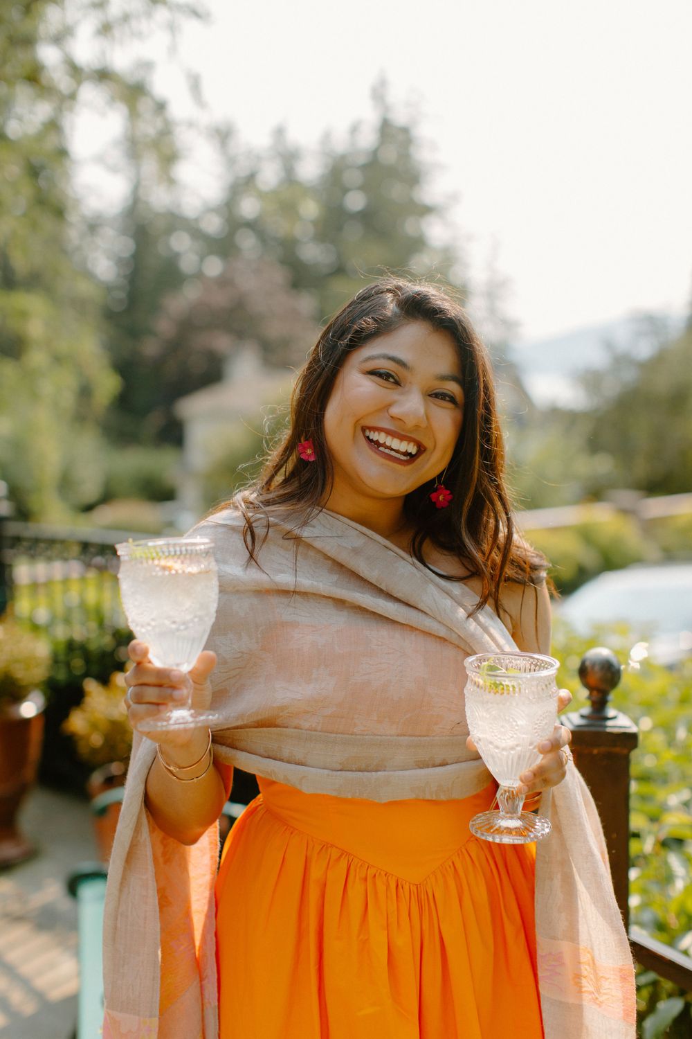 A person wearing an orange skirt and beige wrap holds two cocktail glasses while smiling in a sunny garden setting.