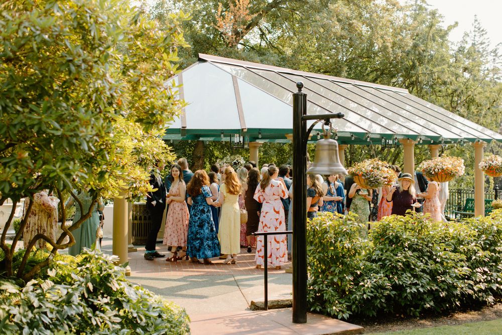 Wedding guests gather under an elegant white pergola surrounded by lush garden greenery on a sunny afternoon.
