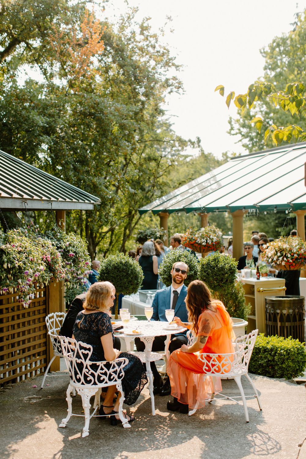 People dining outdoors at an elegant garden cafe with white metal chairs under green awnings and lush landscaping.