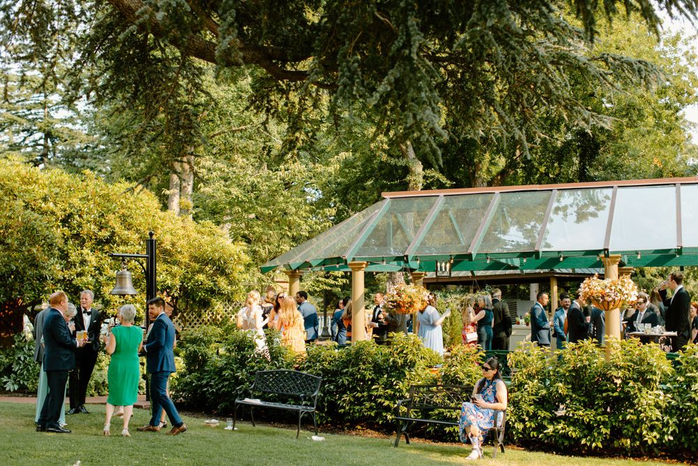 Outdoor garden wedding reception with guests mingling on a lawn near a glass greenhouse structure.