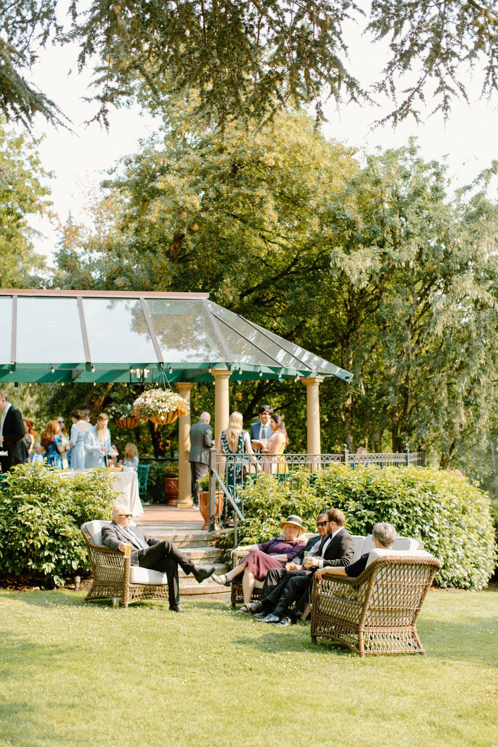 Guests relax in wicker chairs under a covered gazebo in a lush garden setting during an outdoor event.