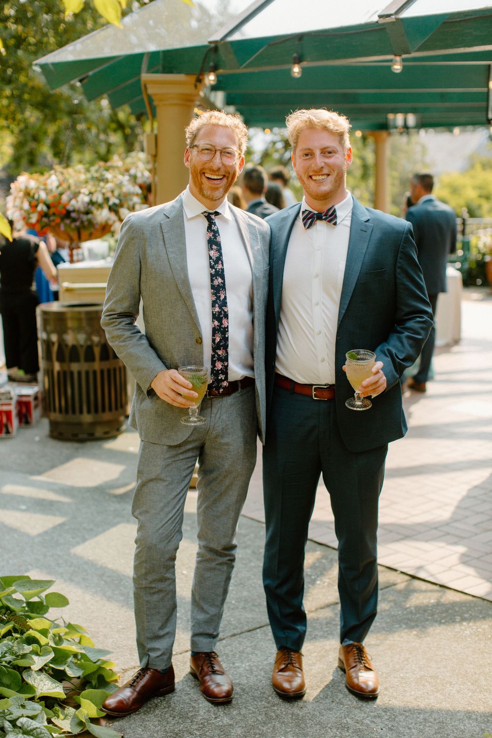 Two well-dressed guests enjoying drinks at an outdoor garden party under a green awning with floral decorations.