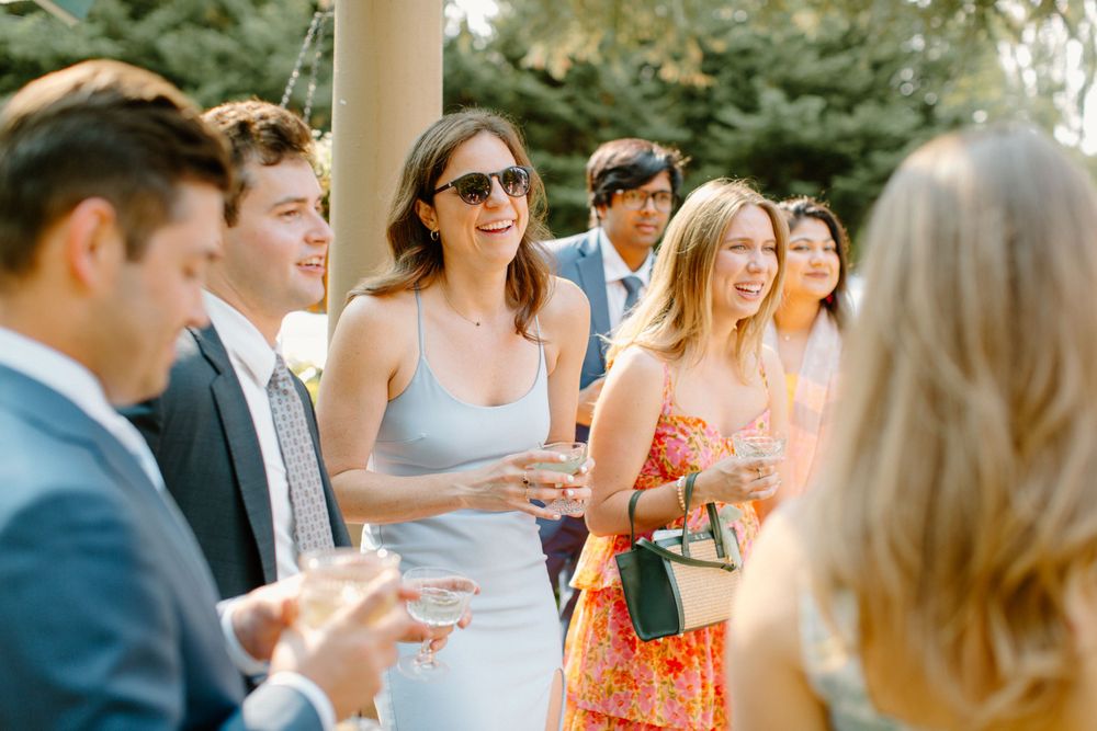 A group of well-dressed guests mingle and chat during an outdoor social gathering while holding beverages.