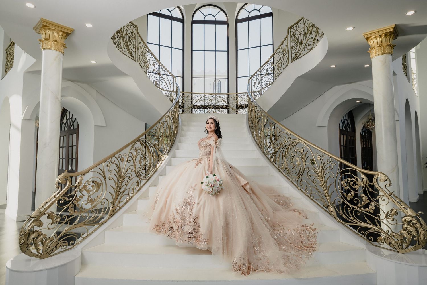 Elegant champagne-colored quince dress with long train photographed on grand staircase with ornate gold railings and arched windows.