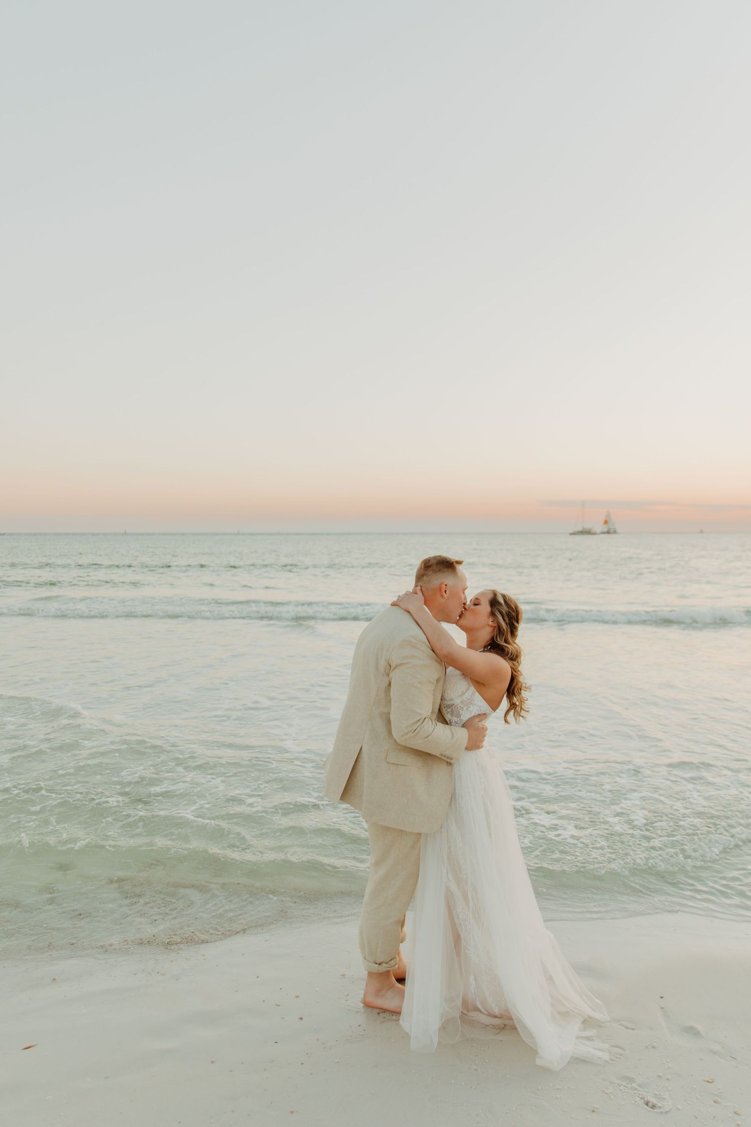 A couple shares a romantic kiss at sunset on a serene beach with soft waves and pastel sky in the background in Panama City Beach, Florida.