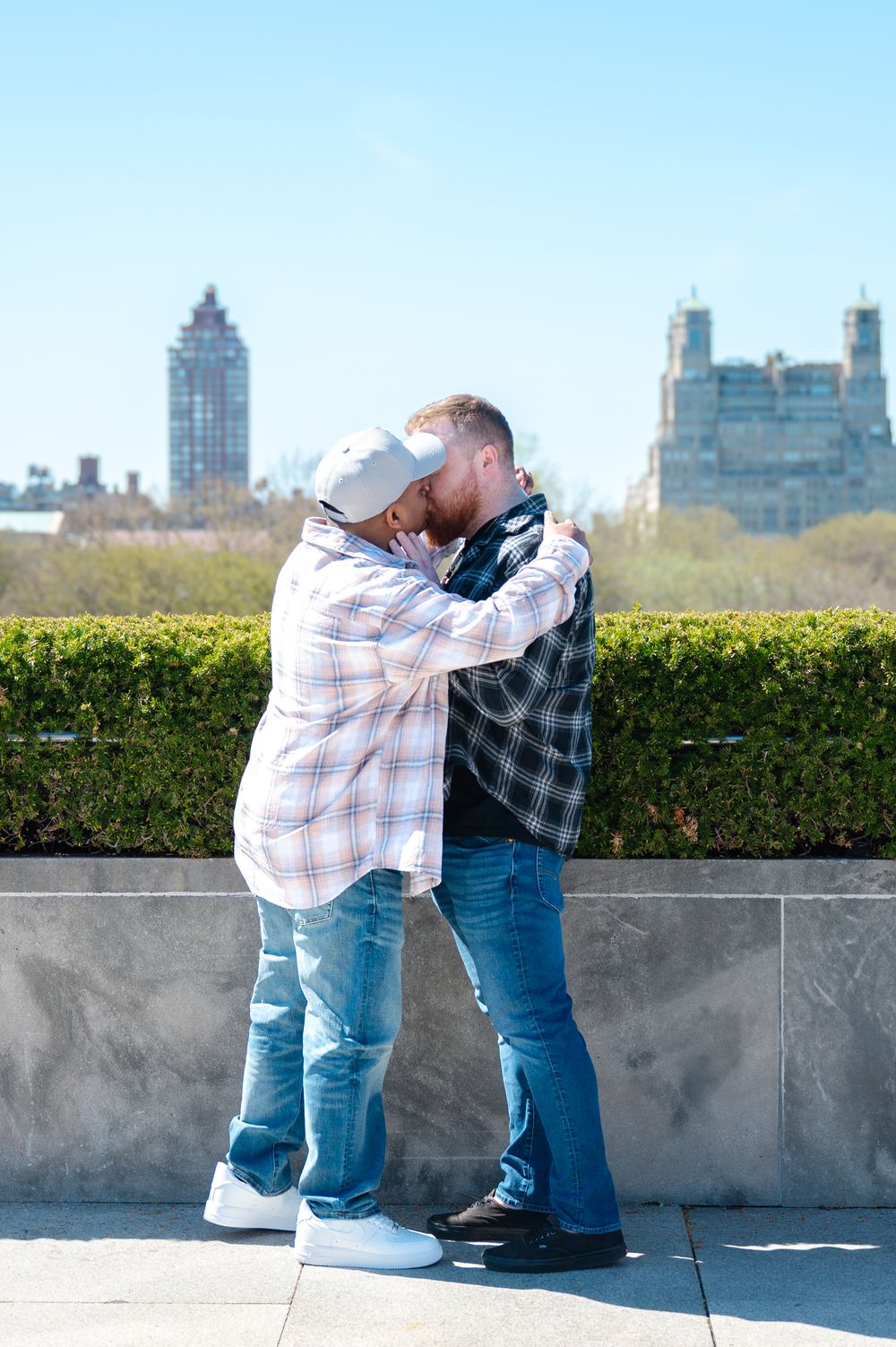 Two men kissing against the backdrop of city buildings and a manicured hedge at a rooftop location after LGBTQ+ proposal at the Met