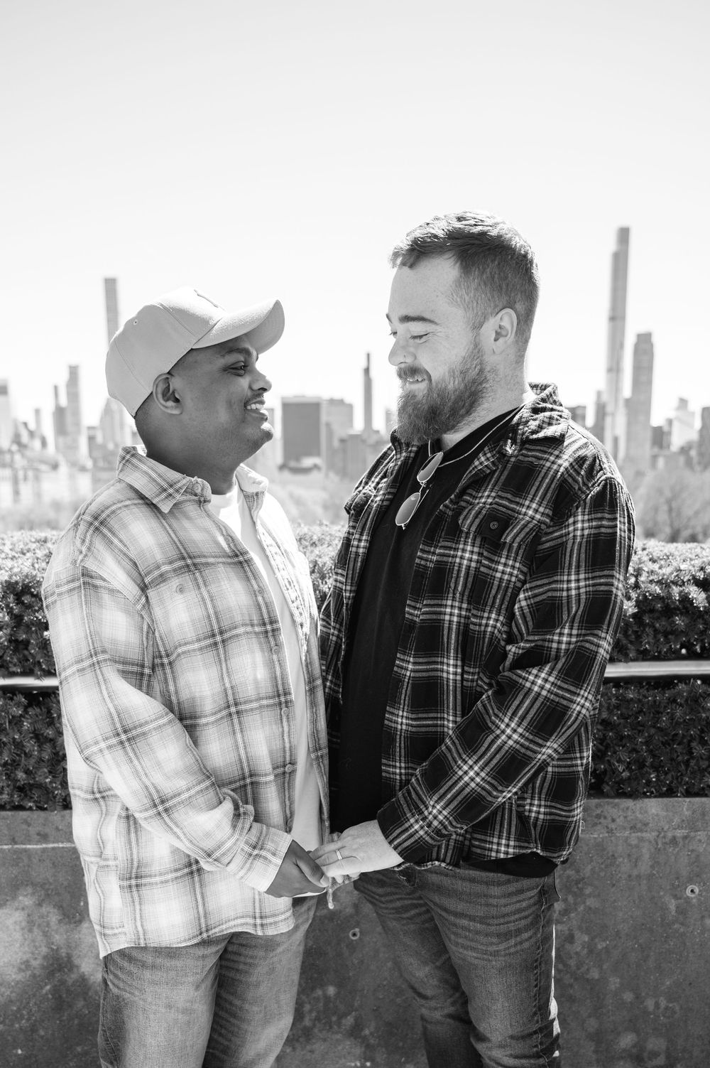 Two men in plaid shirts share a candid moment together with the city skyline visible in the background, black and white engagement photo