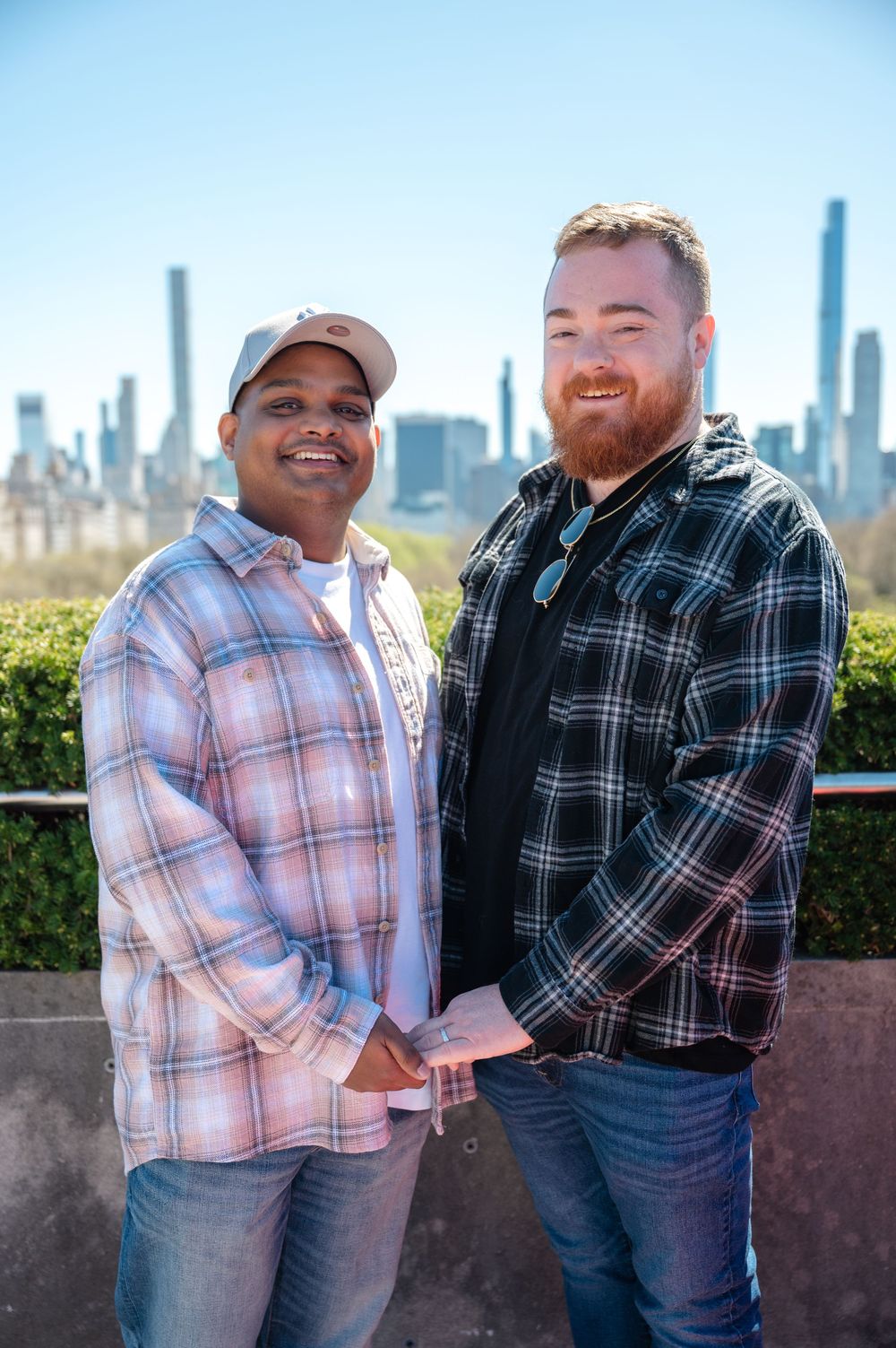 Two men in plaid flannel shirts smile while posing together against a cityscape backdrop on a sunny day after proposal at the Met Museum