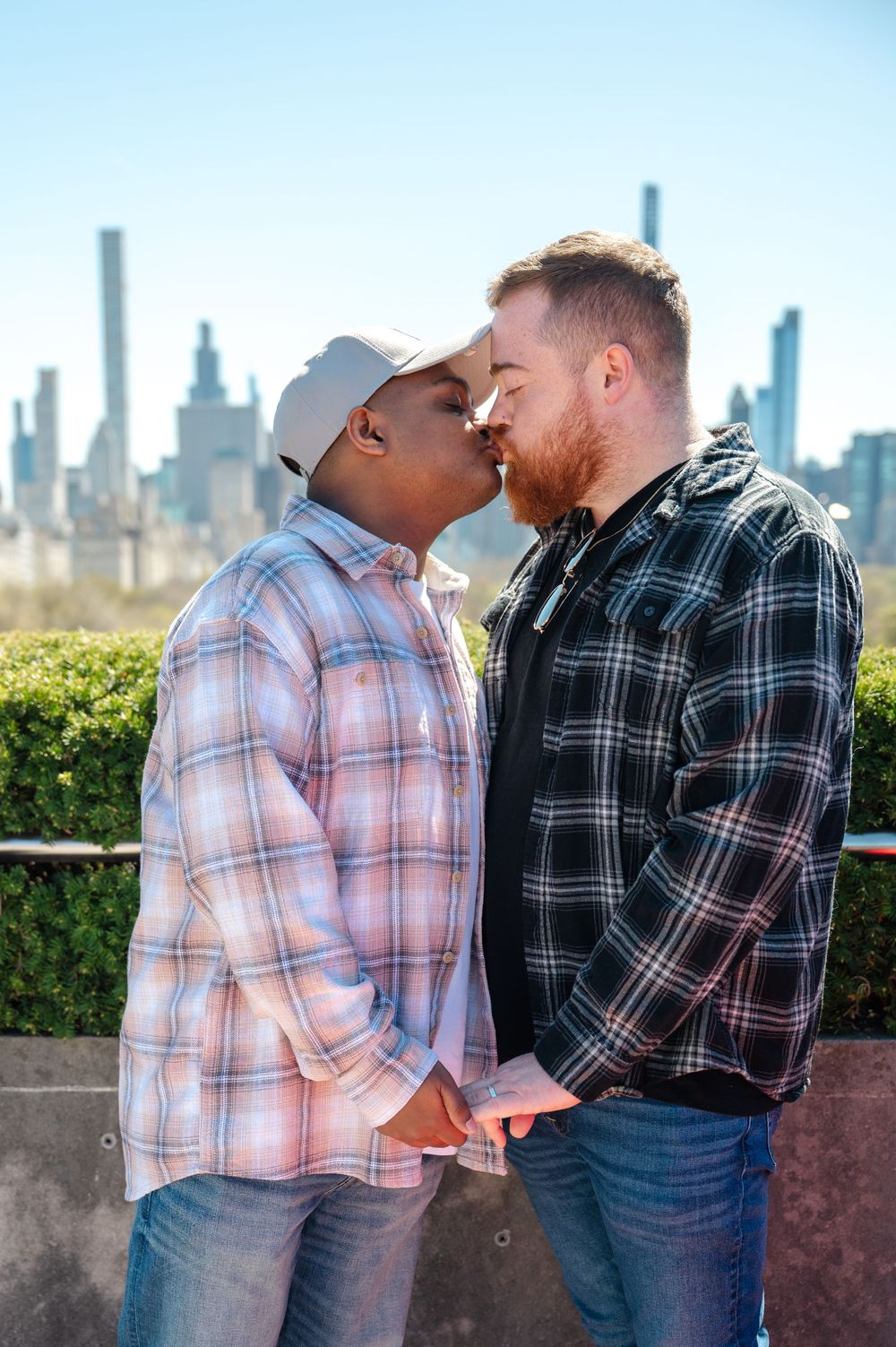 A loving same-sex couple in plaid shirts share an intimate moment with the New York City skyline in the background during engagement session