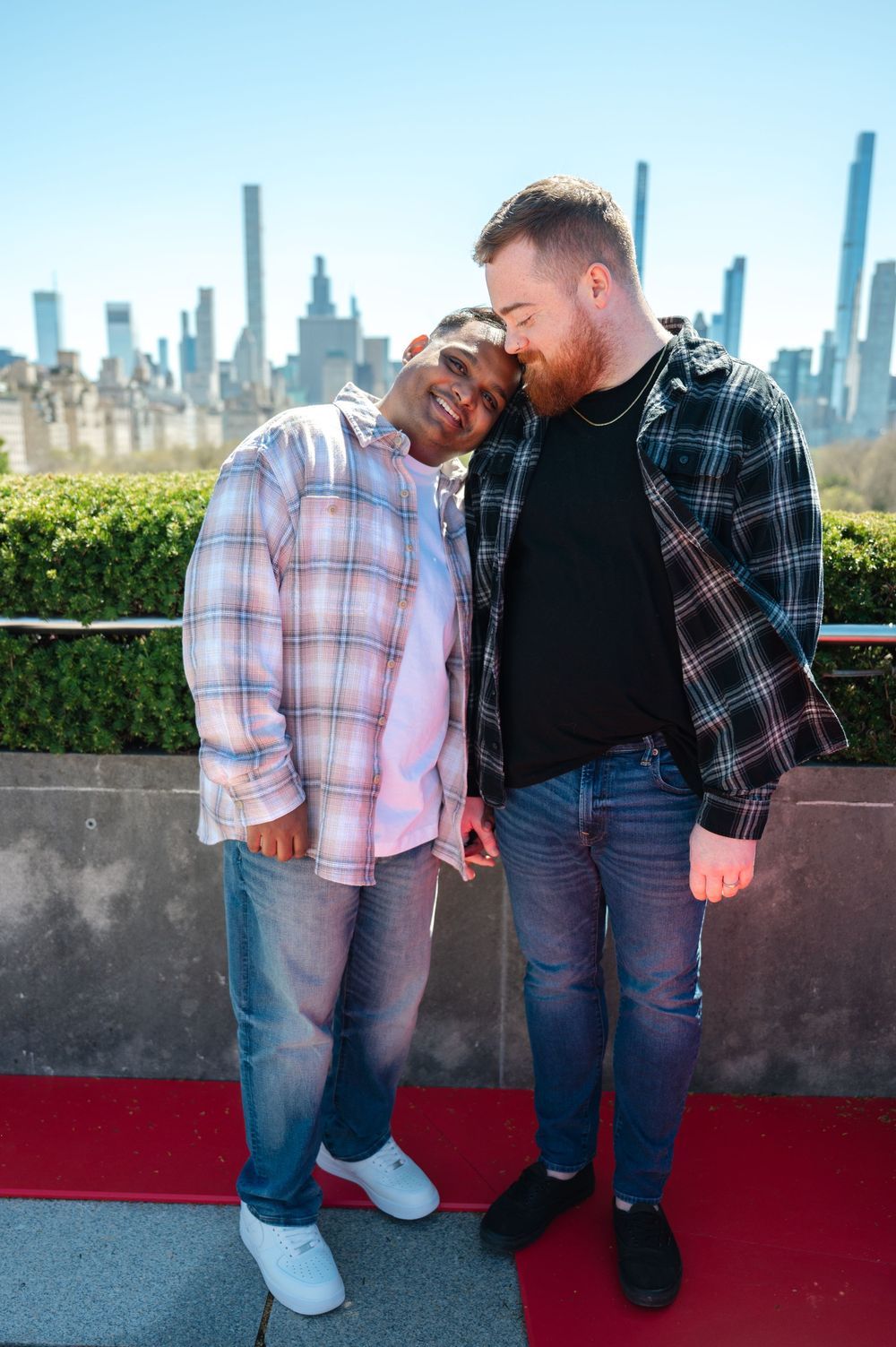 Two men in casual plaid shirts embrace tenderly on a rooftop terrace with Manhattan skyscrapers in the background after same-sex proposal
