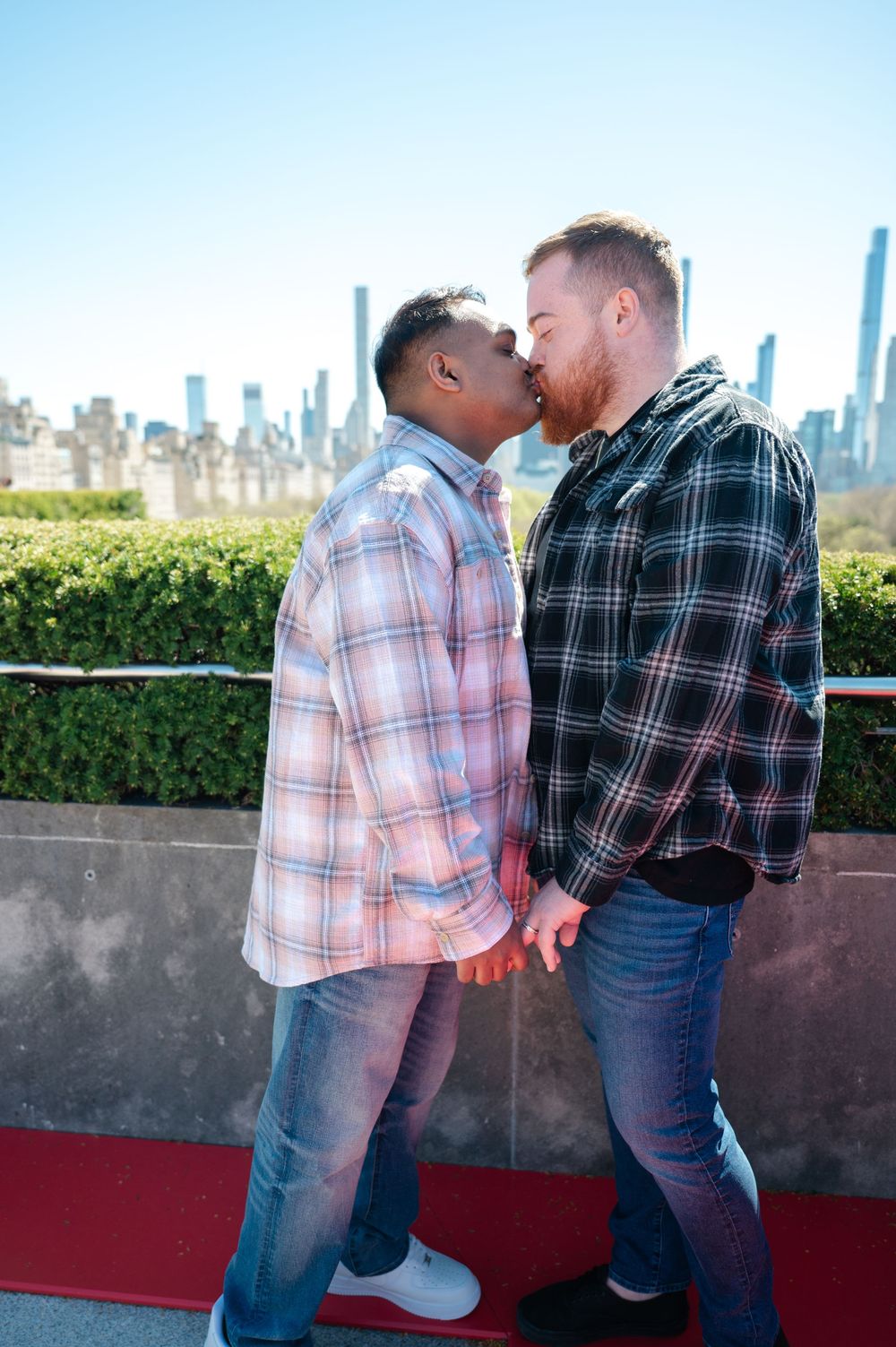 A couple shares an intimate moment overlooking a city skyline while wearing casual plaid shirts after their same-sex engagement at the Met