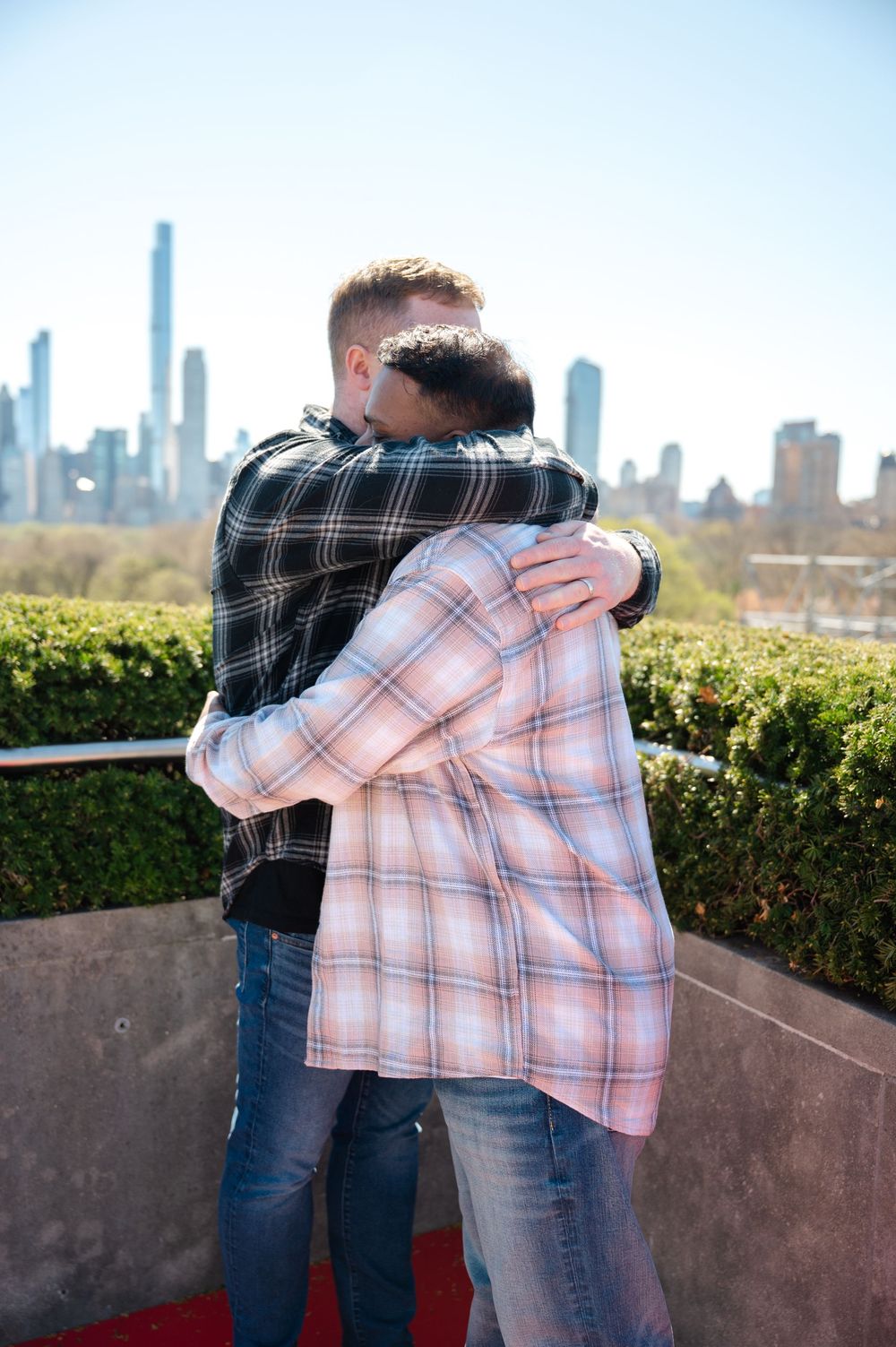 Two newly engaged men in plaid shirts embrace while looking at city skyline from rooftop garden on sunny day at the Met Museum