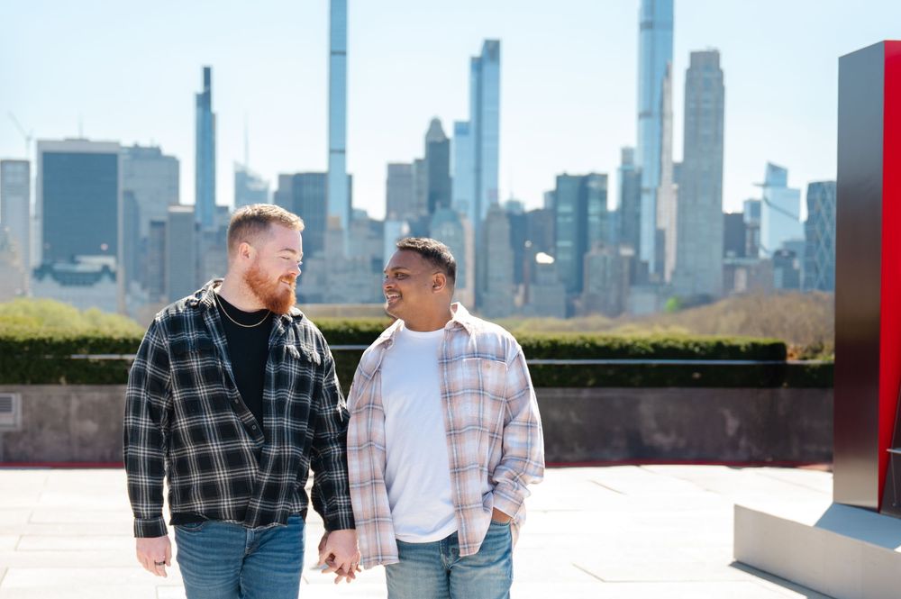 Two men in casual shirts holding hands and smiling with the Manhattan skyline in the background at the Met rooftop after their proposal
